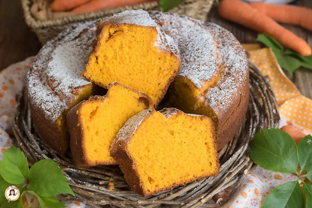 Una soffice torta di carote, cosparsa di zucchero a velo, servita in un cestino di vimini. Le fette mostrano l'interno arancione e invitante.