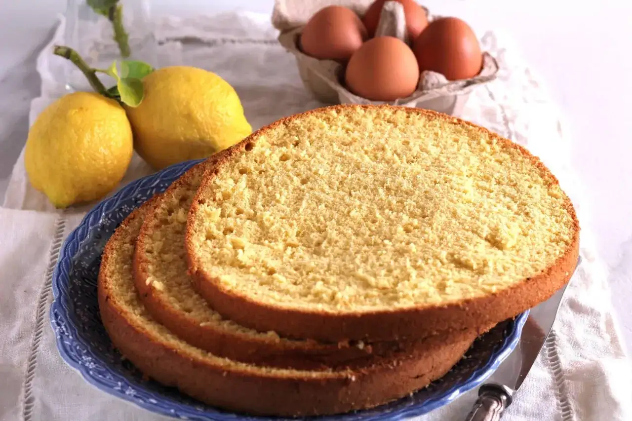 Fette di pan di spagna soffice su un piatto blu, con limoni e uova sullo sfondo, ingredienti per una torta deliziosa.