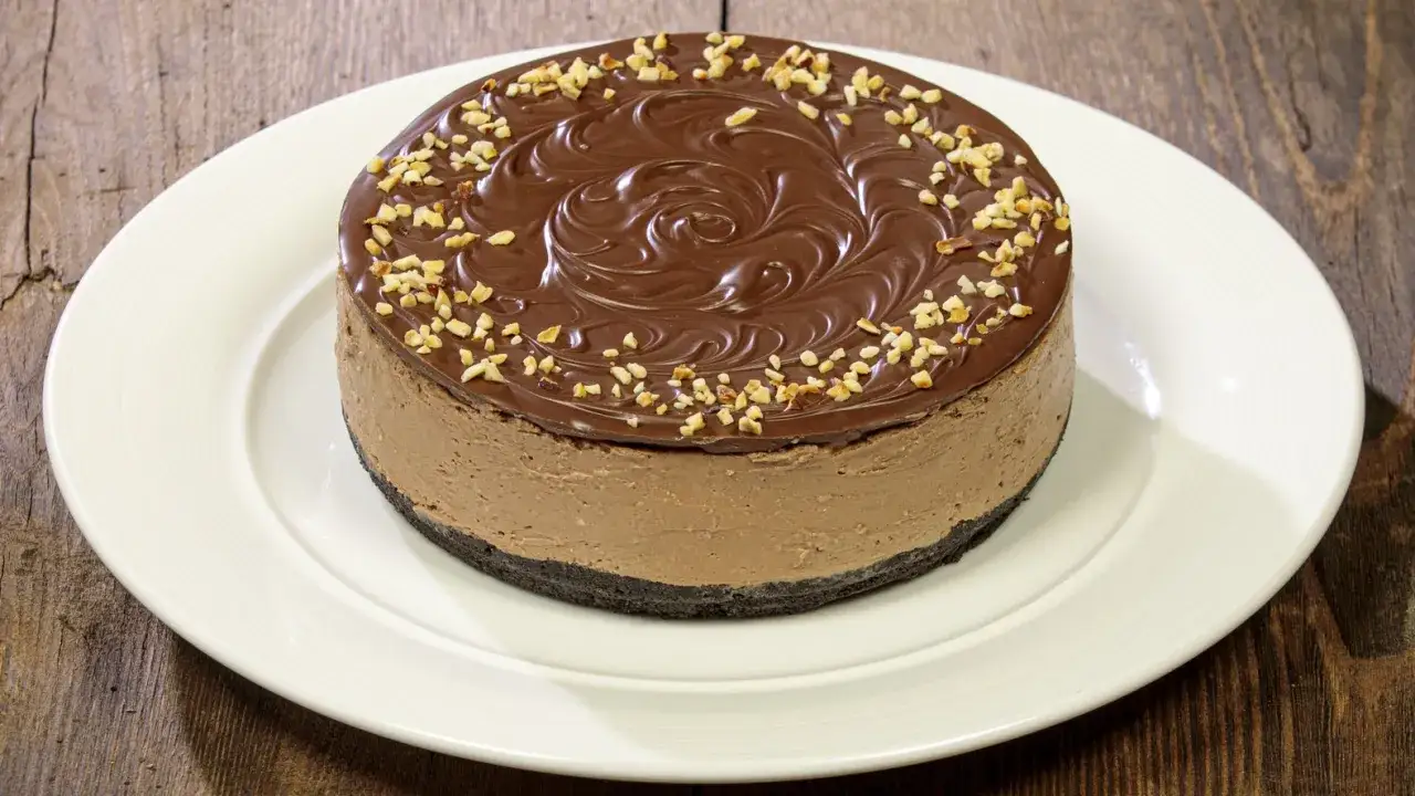 Cheesecake alla Nutella con glassa al cioccolato e nocciole tritate, su un piatto bianco.