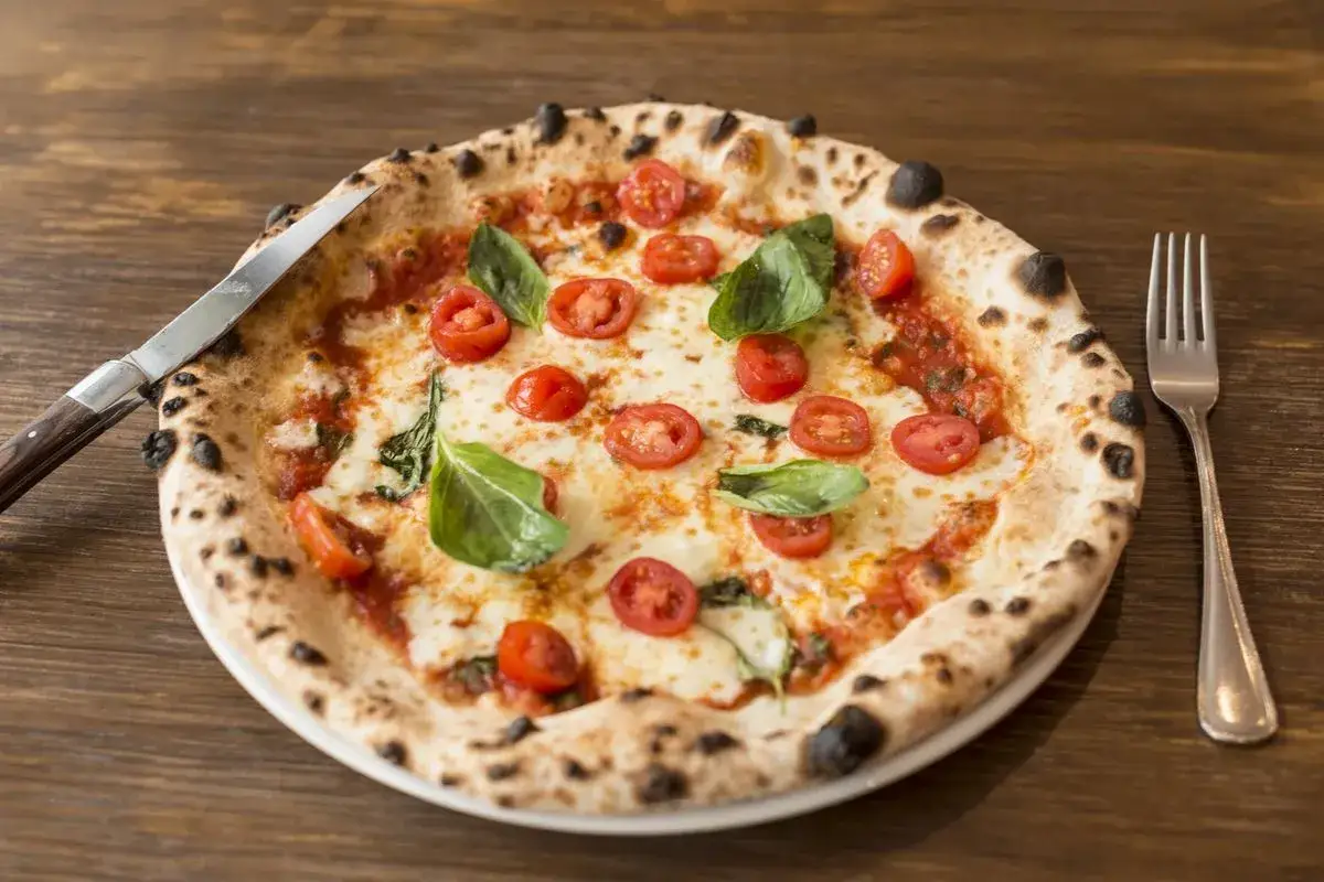 Pizza con lievito di birra, pomodorini freschi, mozzarella filante e basilico profumato, pronta per essere gustata con coltello e forchetta.