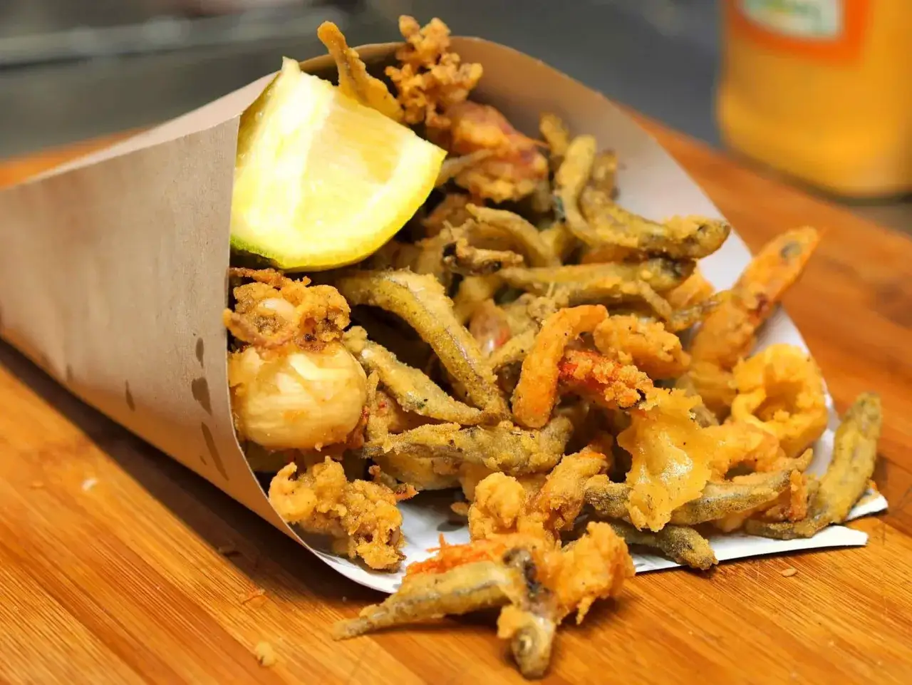 Un cono di carta marrone trabocca di una deliziosa frittura di pesce croccante, con gamberetti, calamari e pesciolini, guarnita con uno spicchio di limone.