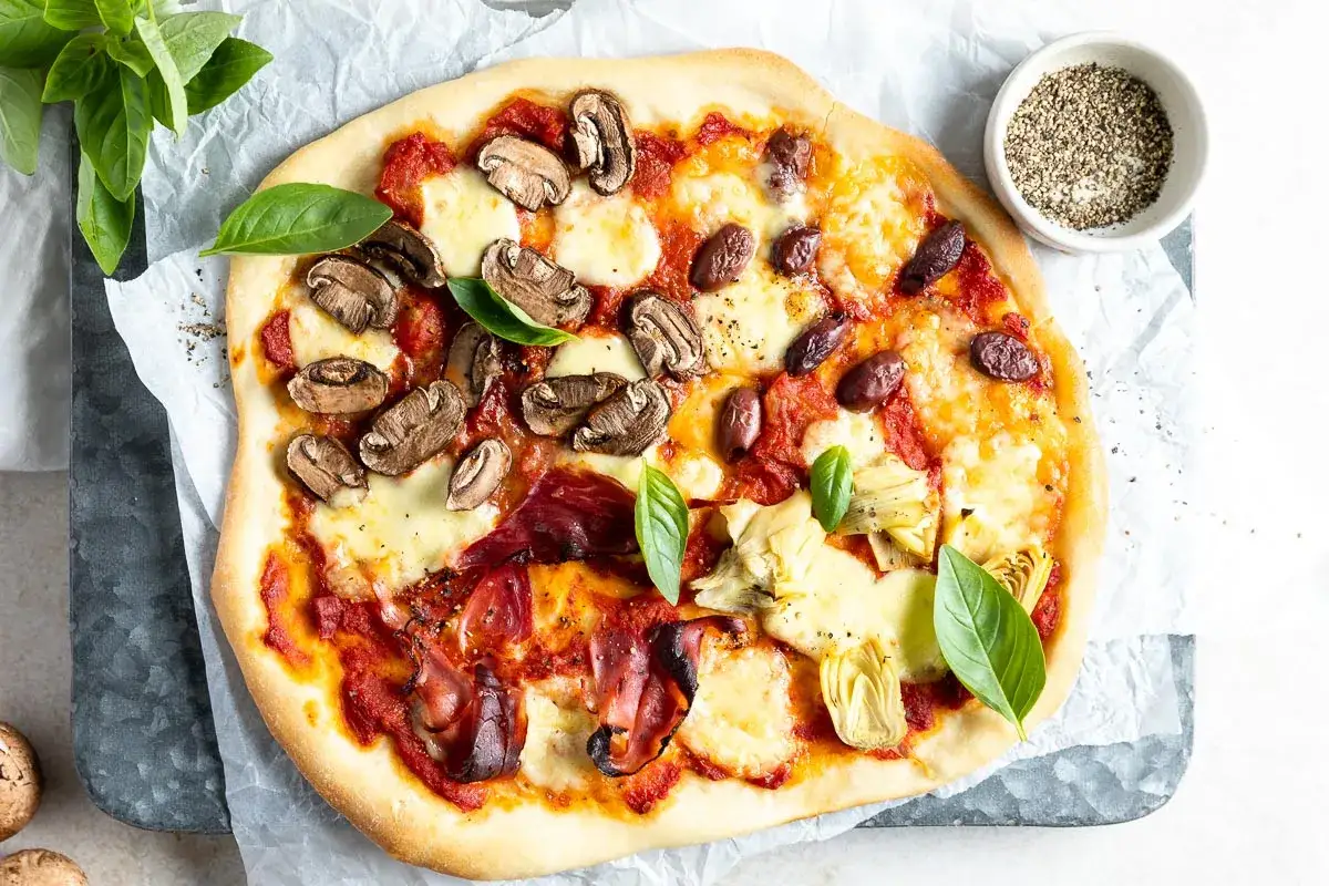 Pizza 4 stagioni con funghi, olive, carciofi e prosciutto crudo, guarnita con basilico fresco.