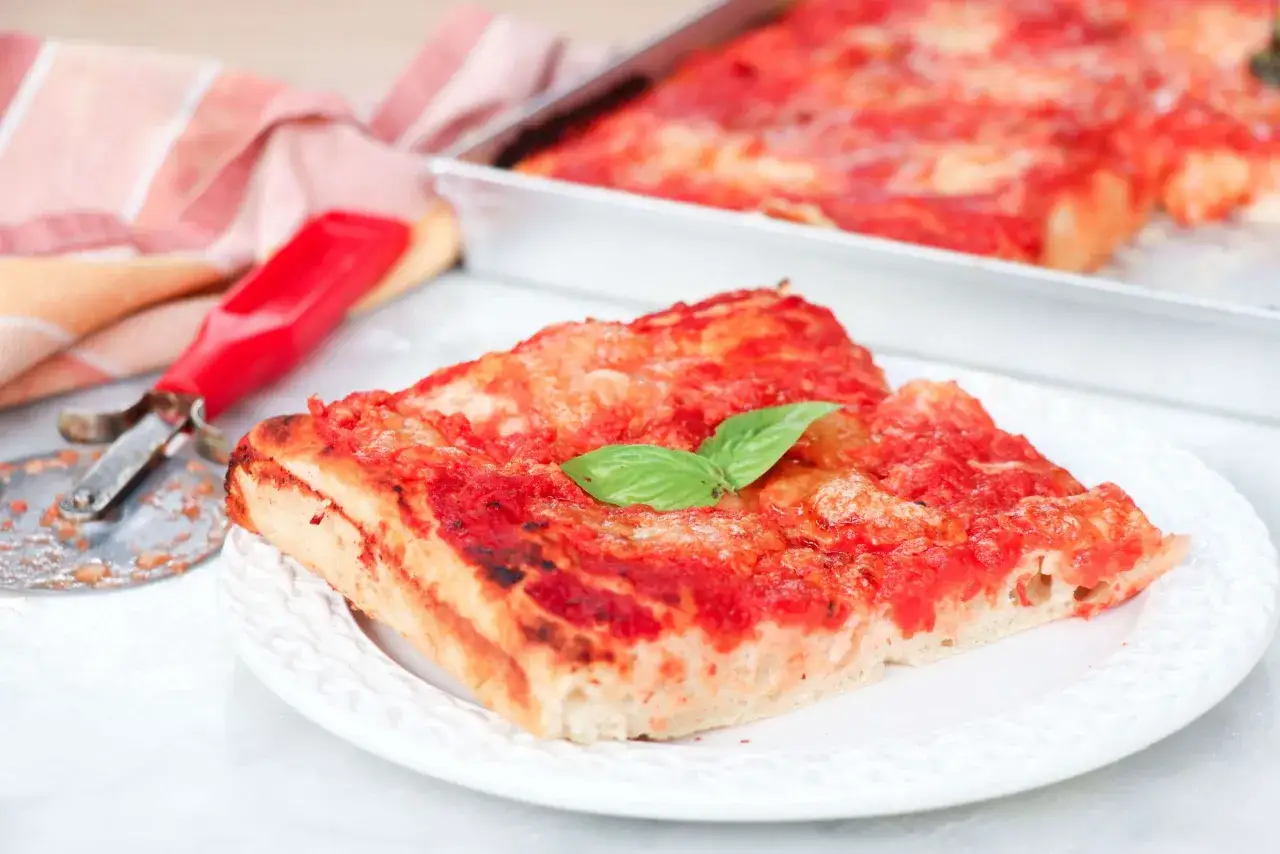 Una fetta di pizza senza glutine, con pomodoro e basilico, servita su un piatto bianco. Accanto, un tagliapizza rosso e un tovagliolo a righe.