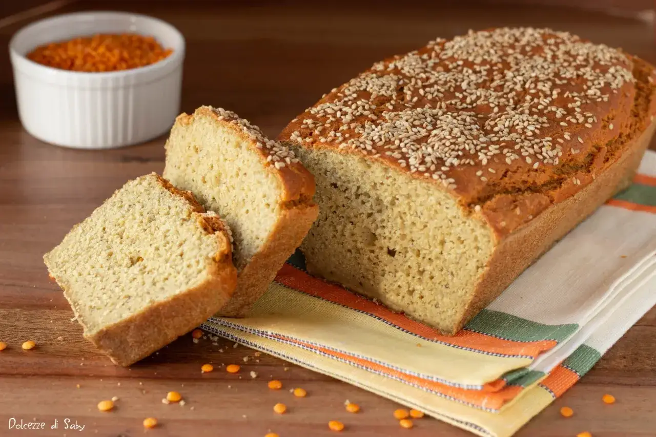 Pane di lenticchie dorato e soffice, cosparso di semi di sesamo, con due fette tagliate. Sullo sfondo, una ciotola di lenticchie rosse.