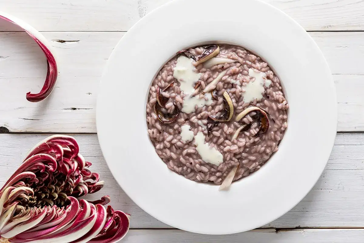 Un cremoso risotto al radicchio, con foglie rosse e bianche di radicchio fresco a lato, su un tavolo di legno bianco.