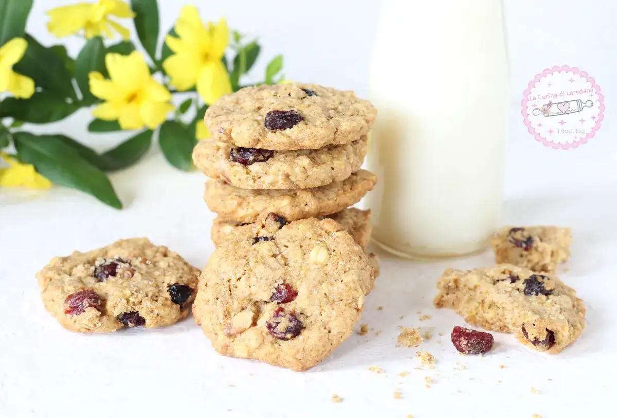 Biscotti senza zucchero con mirtilli rossi e avena, perfetti per una colazione sana. Accanto, un bicchiere di latte e fiori gialli.