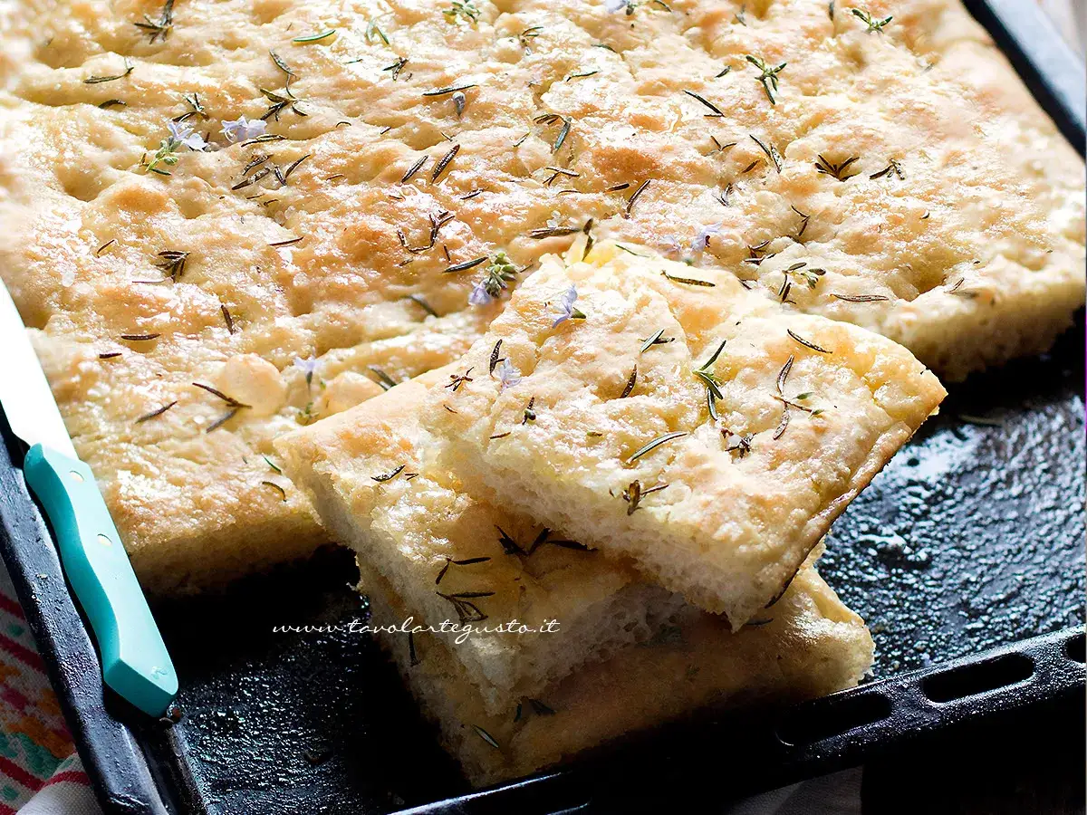 Focaccia senza lievito dorata e profumata, guarnita con rosmarino e piccoli fiori viola, pronta per essere gustata.