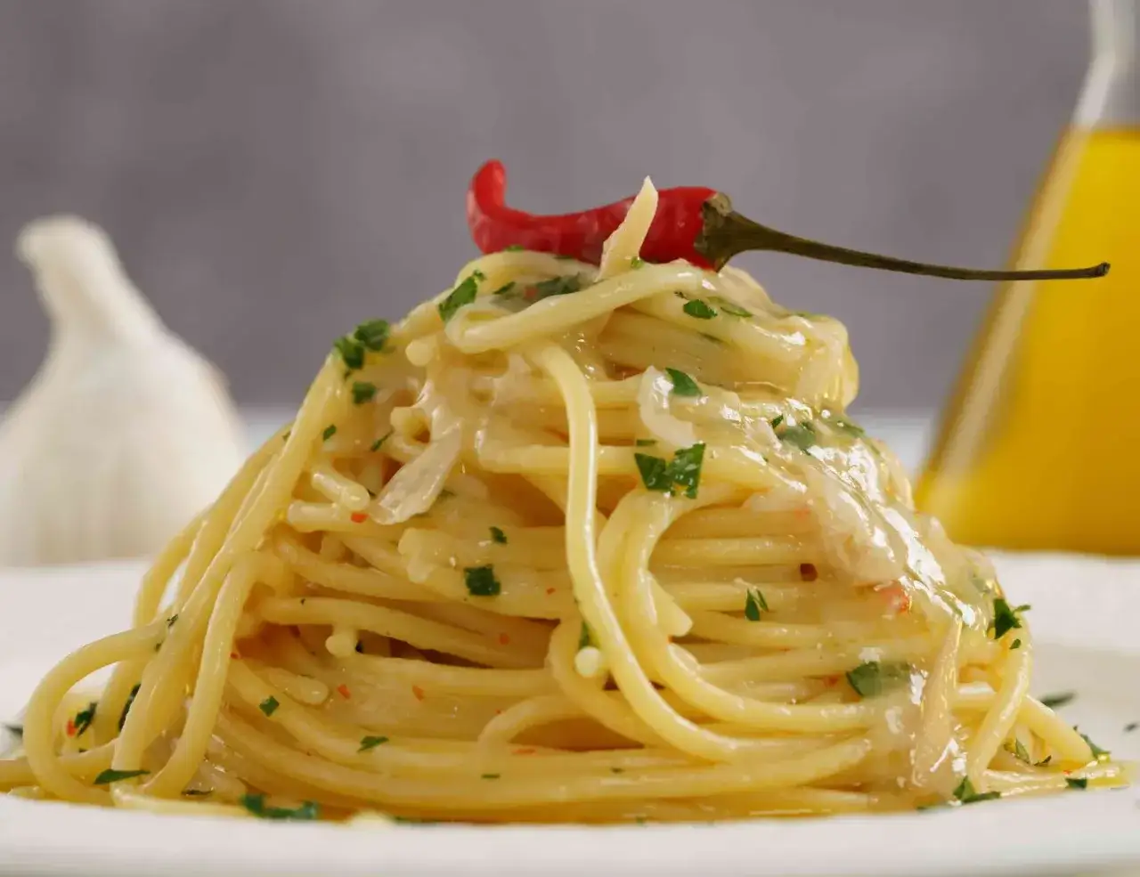 Un piatto invitante di spaghetti aglio olio e peperoncino cremosi, guarnito con un peperoncino rosso e prezzemolo. Sullo sfondo, aglio e olio d'oliva.