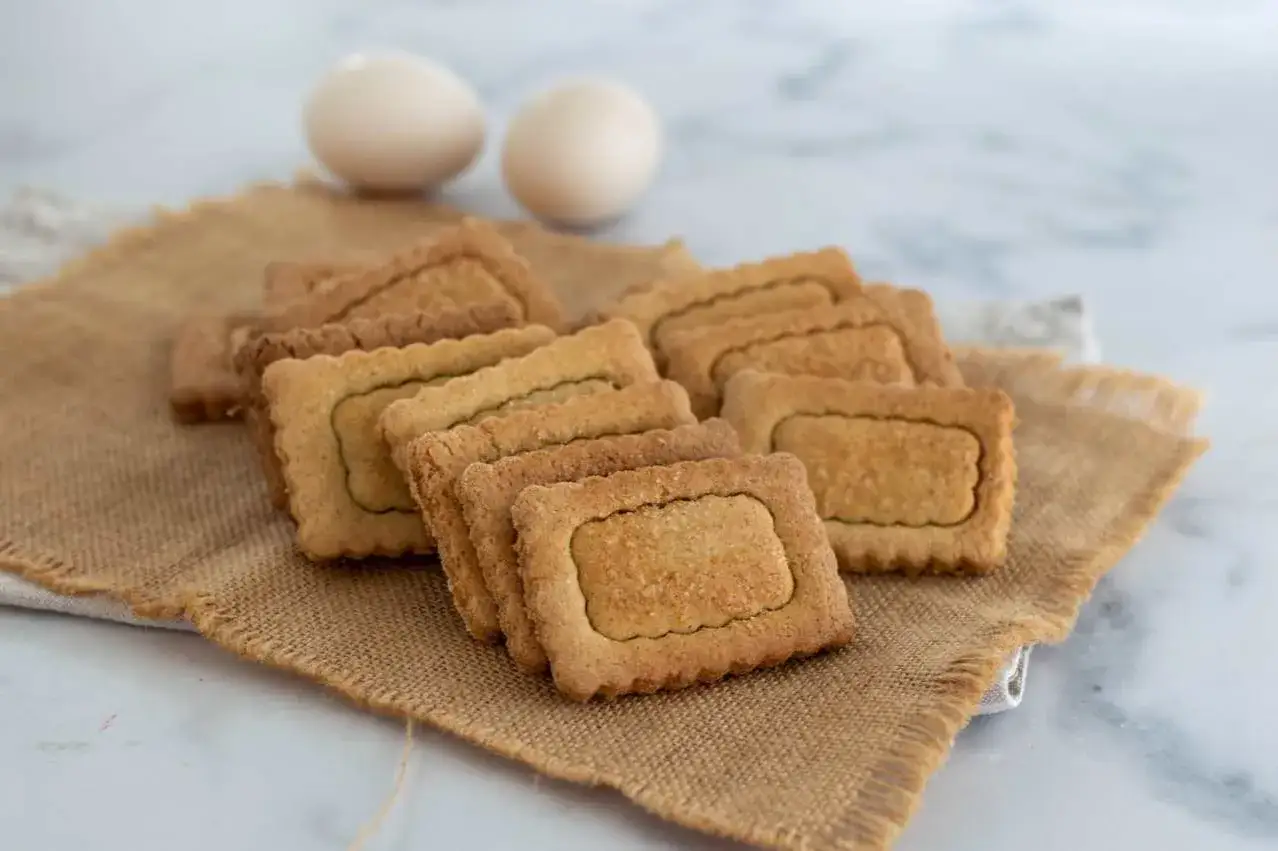 Una pila di biscotti senza uova, dorati e croccanti, su una tela di juta. Due uova in lontananza, un invito a gustare questi deliziosi biscotti senza uova.