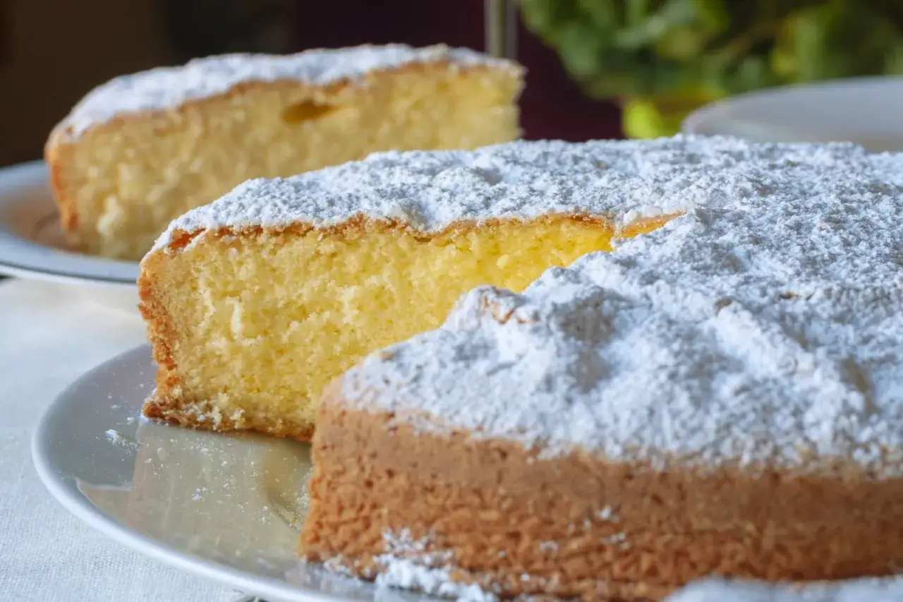 Una fetta di torta paradiso sofficissima, cosparsa di zucchero a velo, su un piatto bianco. Un'altra fetta sfocata sullo sfondo.