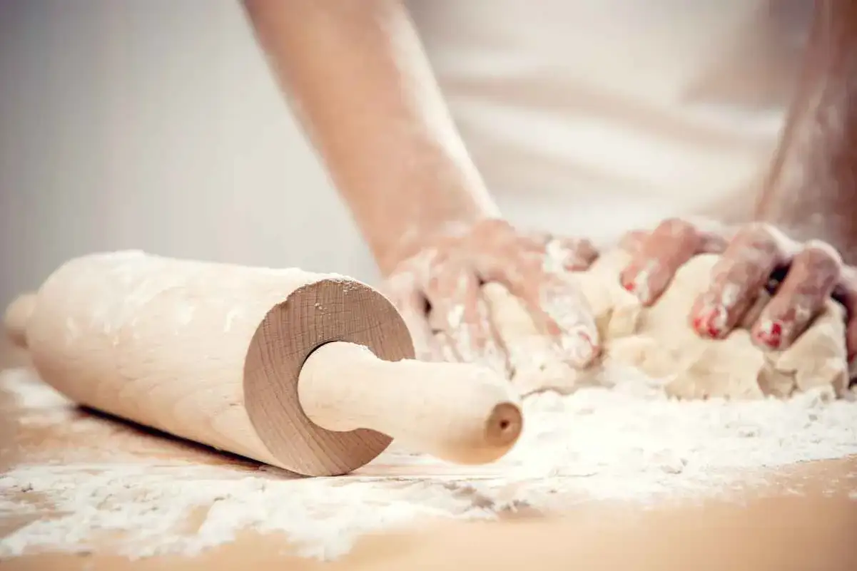 Mani infarinate impastano con un mattarello in primo piano, preparano tagliatelle fatte in casa.