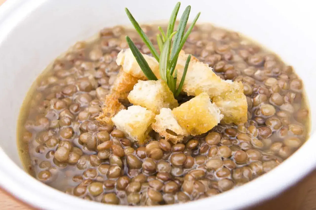 Una ciotola di minestra di lenticchie fumante, guarnita con crostini dorati e un rametto di rosmarino fresco.
