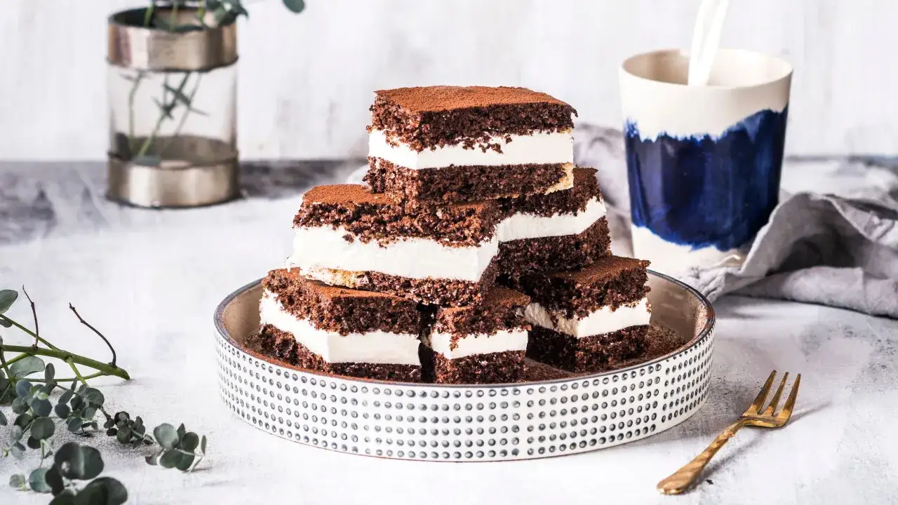 Fette fette di torta all'acqua al cacao con crema bianca, spolverate di cacao, su un piatto decorato. Accanto, una tazza con latte versato e un rametto di eucalipto.