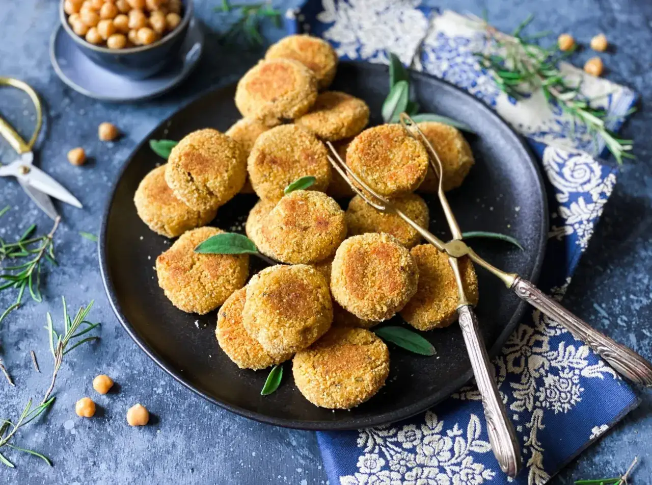 Piatto di polpette di ceci dorate, guarnite con foglie verdi, su sfondo scuro. Vicino, forbici dorate e ciotola di ceci.