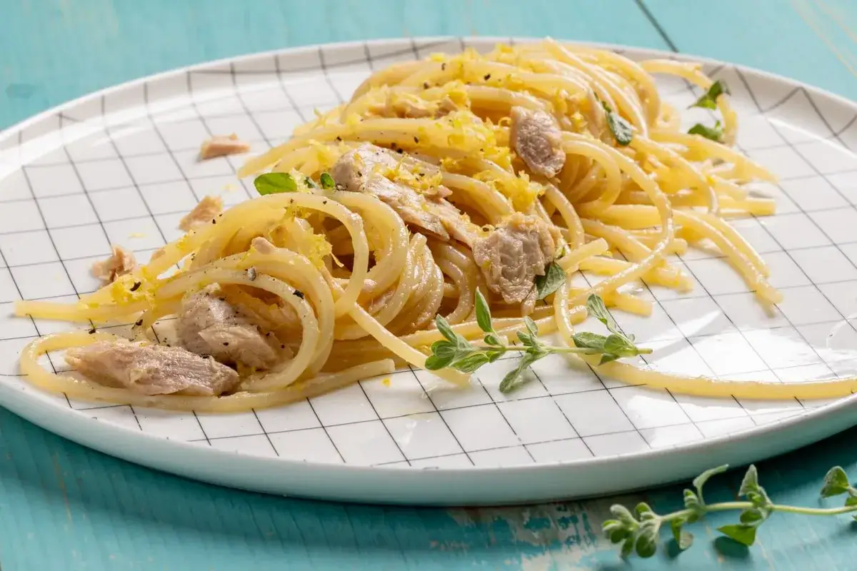 Un piatto di pasta tonno e limone, condita con scorza di limone grattugiata e erbe aromatiche fresche, servita su un piatto a scacchi bianchi.