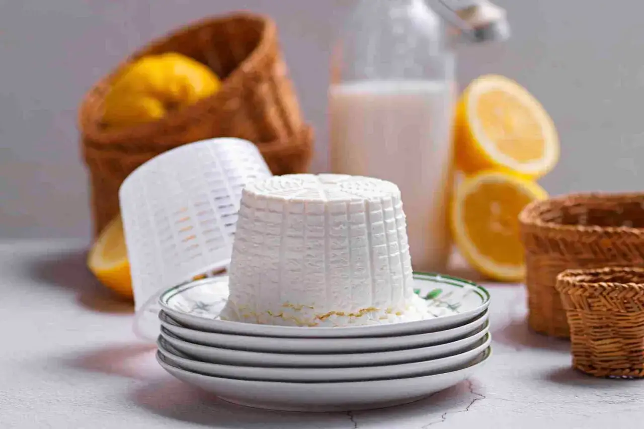 Ricotta fatta in casa appena scolata, con limoni, latte e cestini in primo piano.