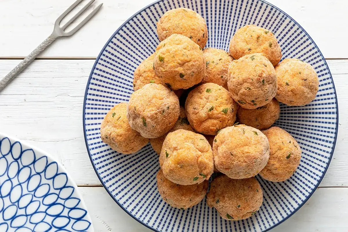 Un piatto pieno di polpette di merluzzo dorate, pronte per essere gustate. Un forchettone metallico attende accanto.