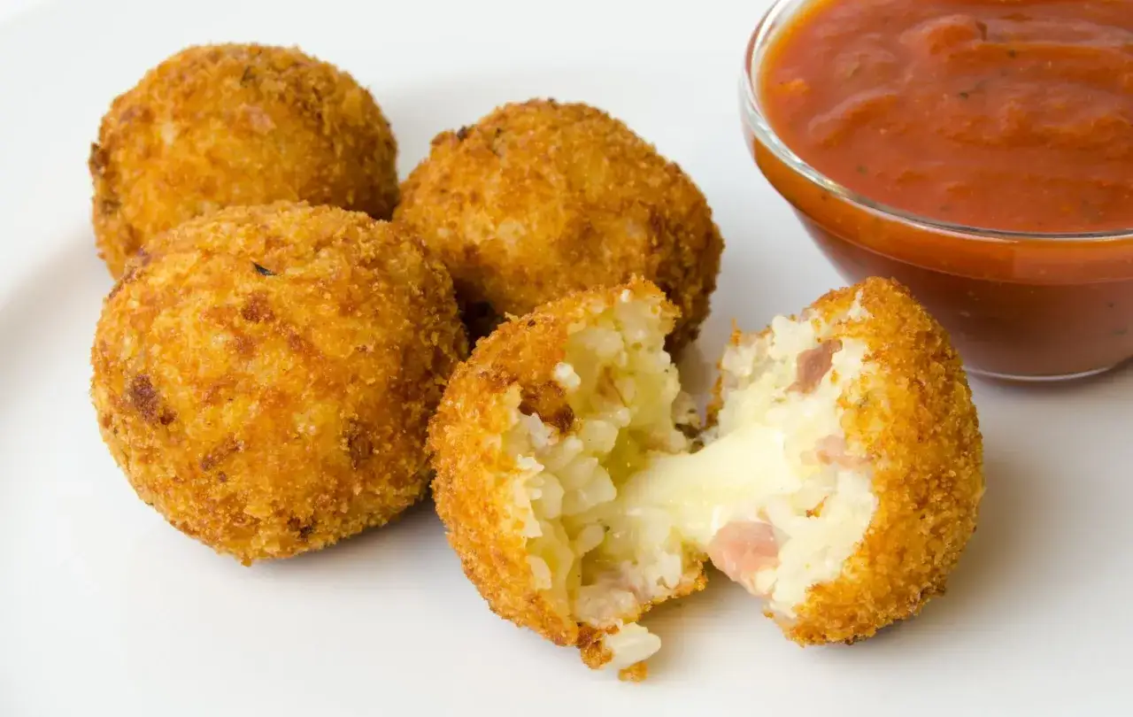 Polpette di riso dorate e croccanti, una aperta rivela un cuore filante di formaggio e prosciutto. Accanto, una ciotola di salsa rossa.