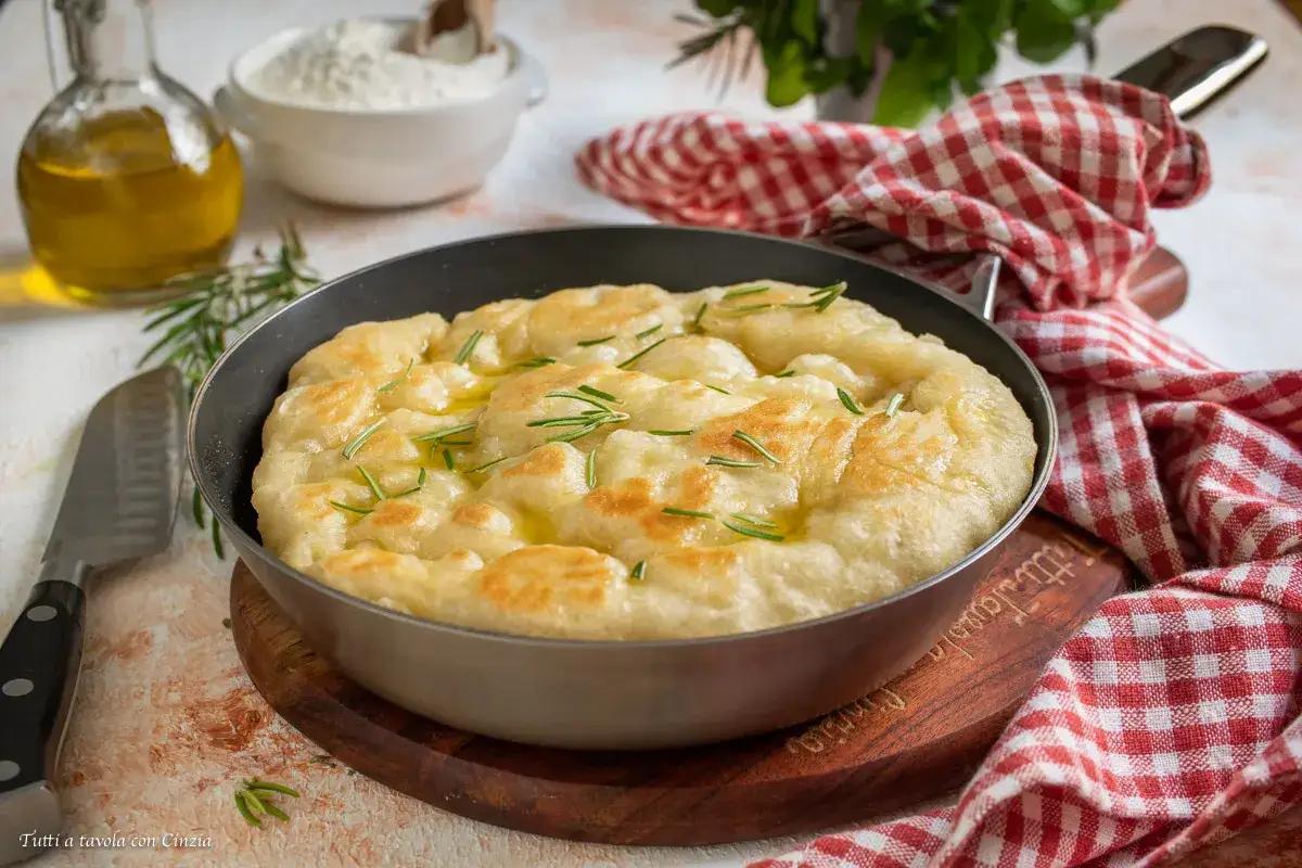 Focaccia in padella dorata e profumata al rosmarino, pronta per essere gustata. Olio d'oliva e farina sullo sfondo.