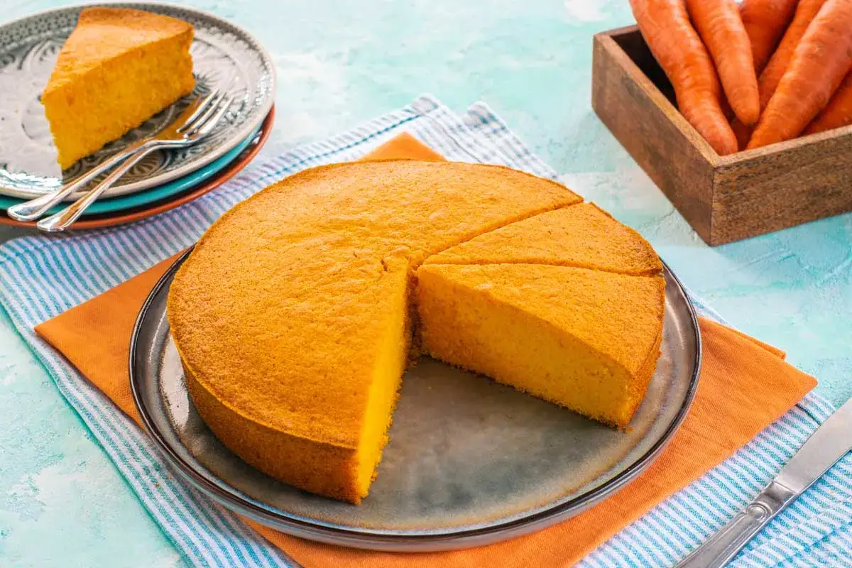 Una deliziosa torta di carote, soffice e dorata, tagliata a fette su un piatto. Accanto, carote fresche in un cesto di legno.