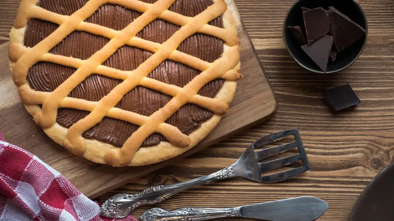Crostata alla Nutella con una griglia di pasta frolla dorata, ripiena di crema al cioccolato. Accanto, pezzi di cioccolato fondente e posate d'argento.