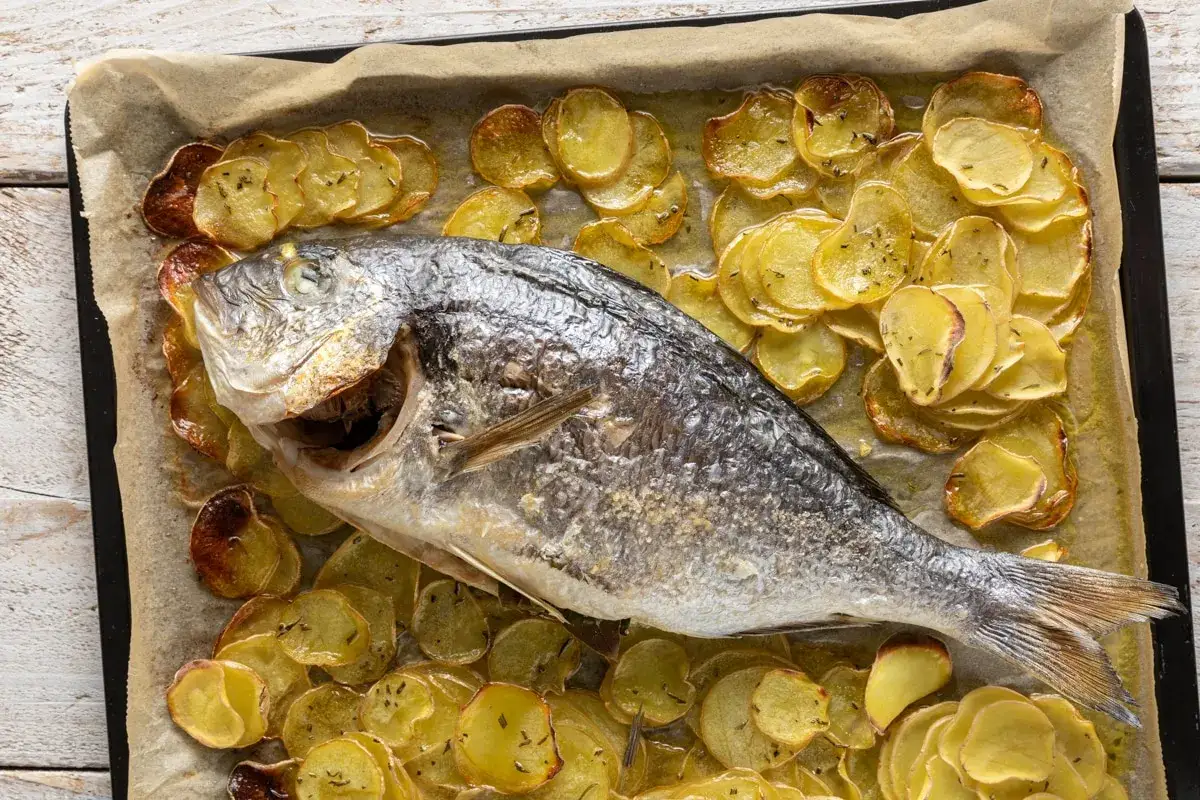 Orata intera cotta in friggitrice ad aria, adagiata su patate a fette dorate e profumate.