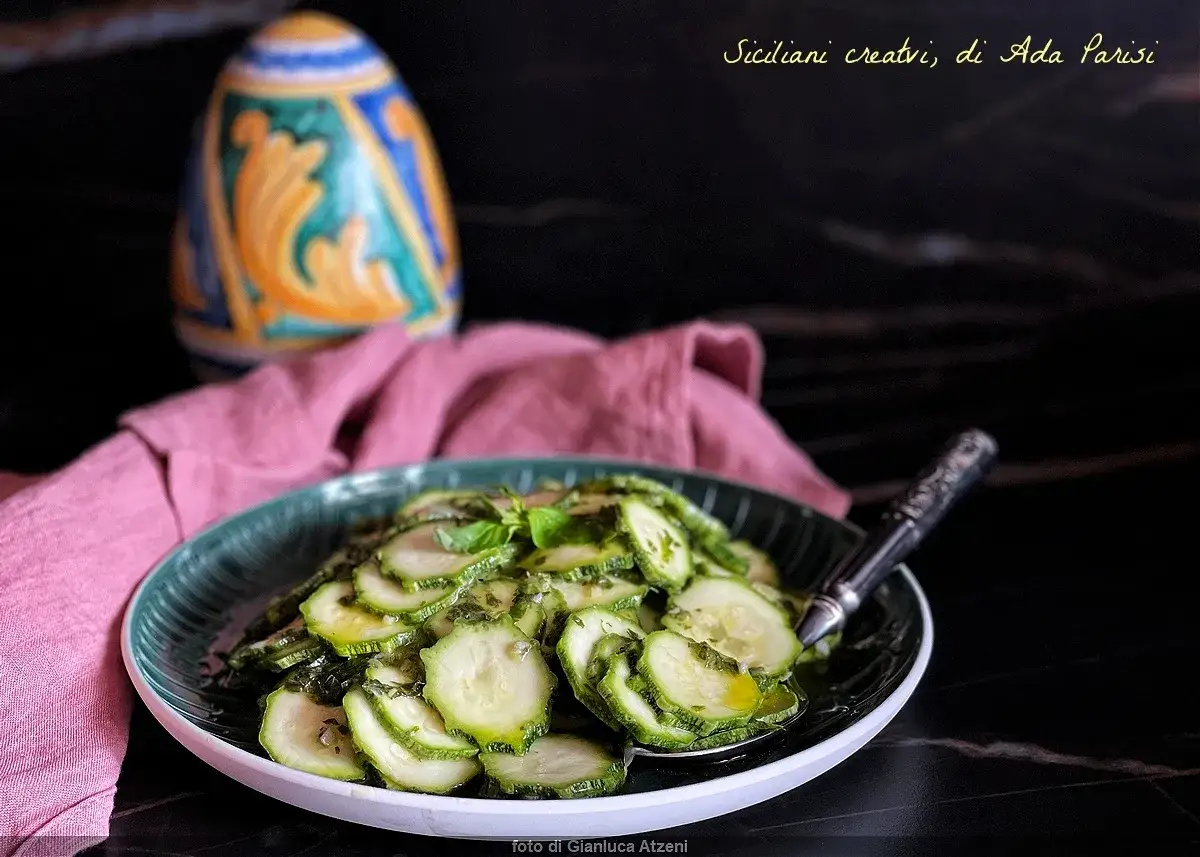 Fette di zucchine marinate crude, condite con erbe aromatiche e olio, servite su un piatto verde.