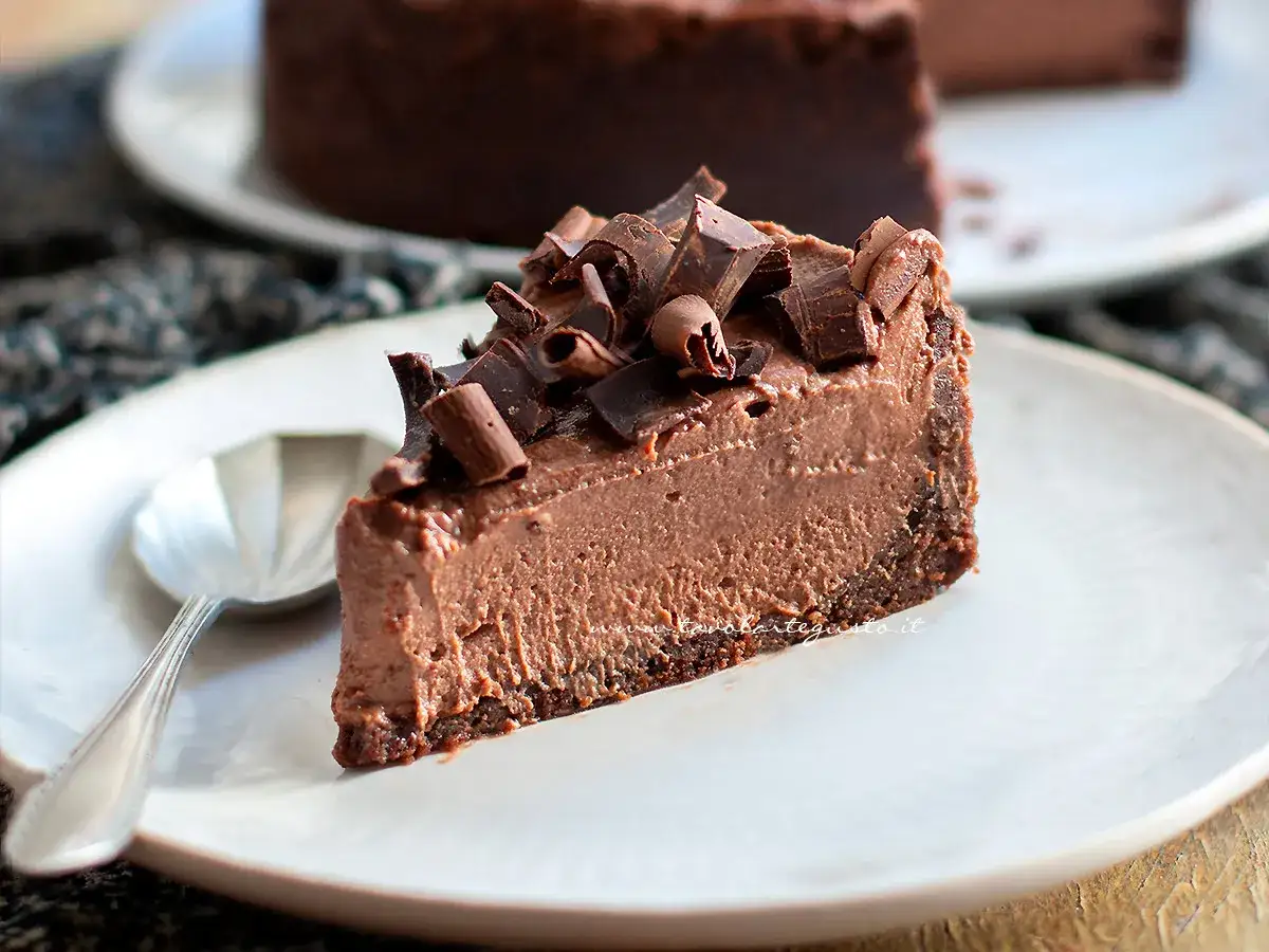 Una fetta di goloso cheesecake al cioccolato, guarnita con riccioli e scaglie di cioccolato fondente, su un piatto bianco con un cucchiaino.