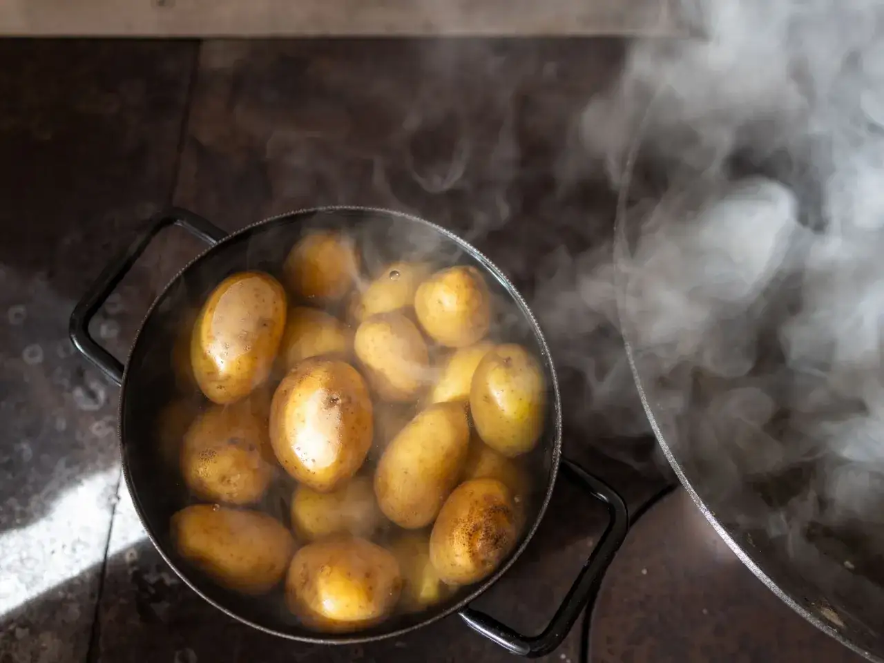Patate intere in pentola che bollono, emanando vapore. Il tempo di cottura patate lesse senza buccia &egrave; quasi terminato.
