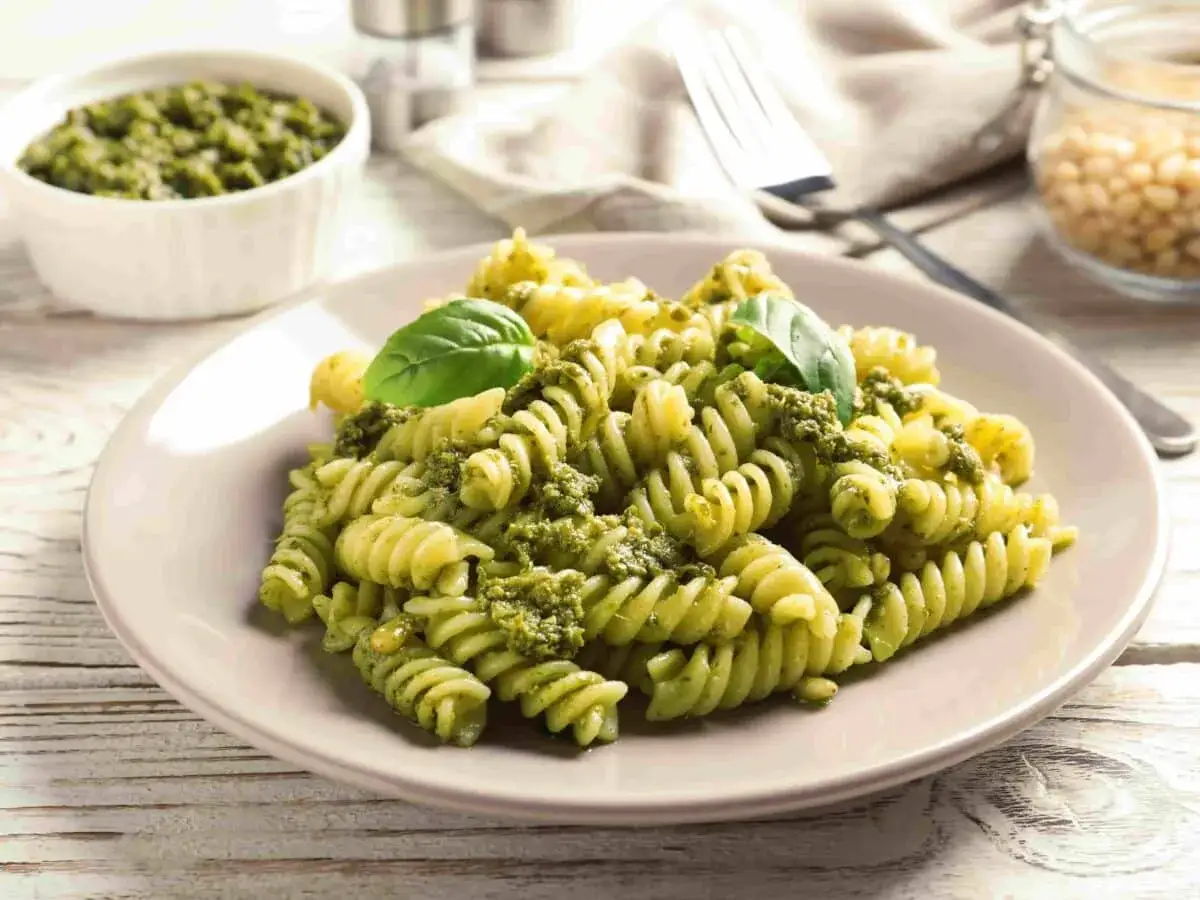 Piatto di fusilli conditi con abbondante pesto alla genovese, guarnito con foglie di basilico fresco. Sullo sfondo, una ciotola di pesto e pinoli.