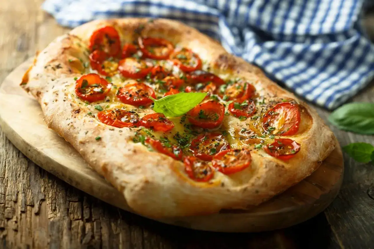Focaccia con lievito istantaneo, condita con pomodorini, erbe aromatiche e basilico fresco, su tagliere di legno.