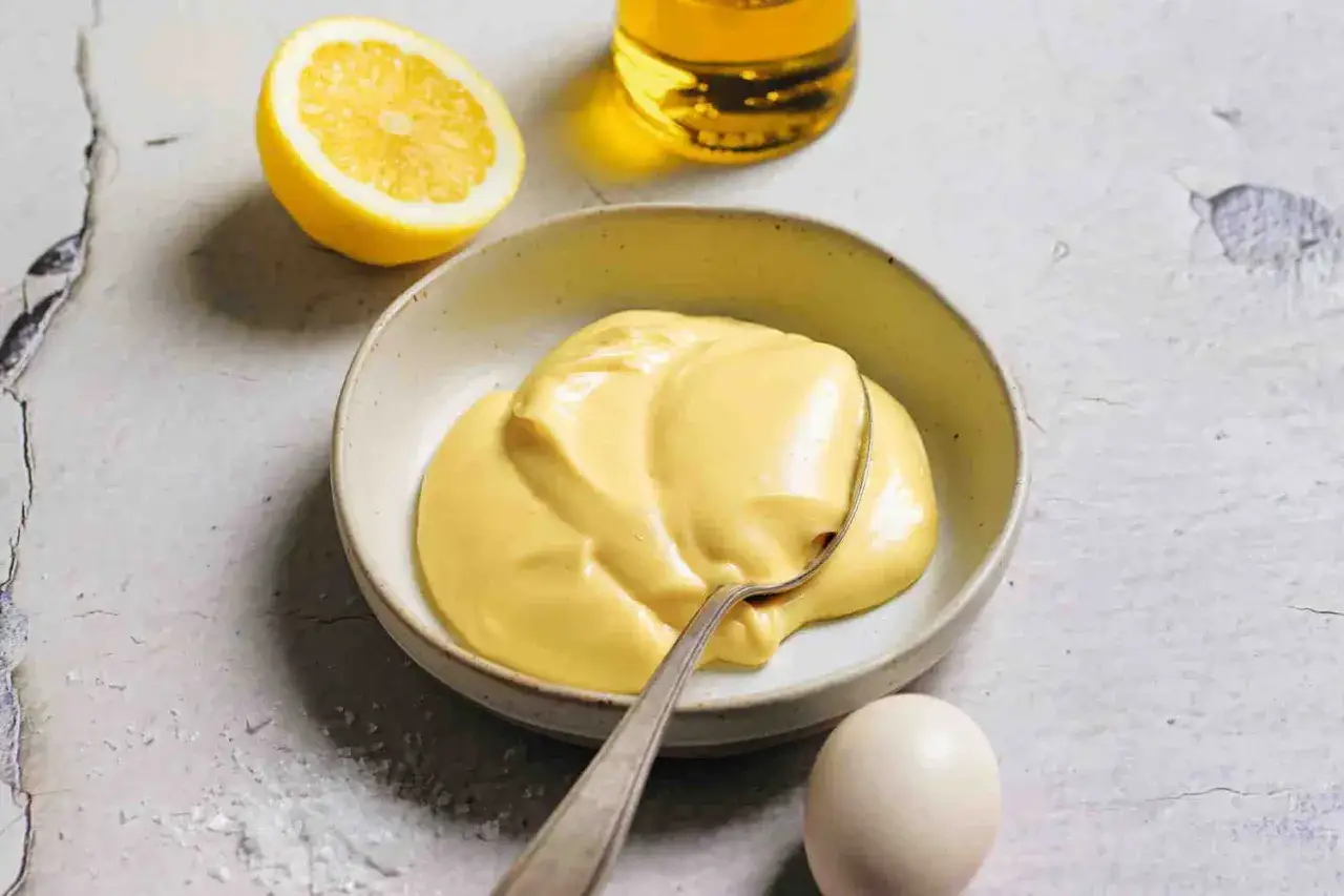 Una ciotola di cremosa maionese fatta in casa, con limone, uovo e olio a fianco.