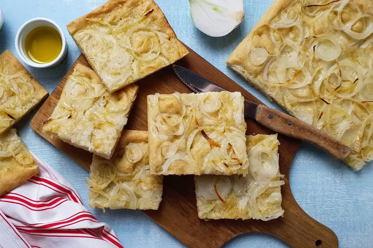Focaccia con cipolle dorate e profumate, tagliata a quadrotti su un tagliere di legno, con un filo d'olio e una cipolla fresca accanto.