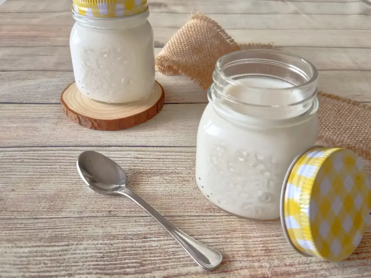 Due vasetti di yogurt fatto in casa, uno con coperchio a quadretti gialli, su un tavolo di legno. Un cucchiaino &egrave; appoggiato accanto.