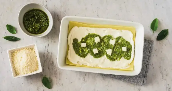 Lasagne al pesto in preparazione: strati di pasta, besciamella, pesto e cubetti di formaggio. Accanto, ciotole con pesto e parmigiano grattugiato.