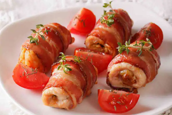 Involtini di pollo avvolti in pancetta croccante, guarniti con timo fresco e serviti con spicchi di pomodoro.