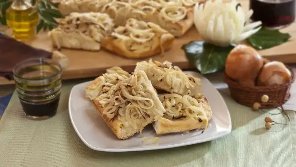 Focaccia con cipolle dorate, fragrante e invitante, servita su un piatto bianco. Sullo sfondo, ingredienti freschi e una bottiglia d'olio.