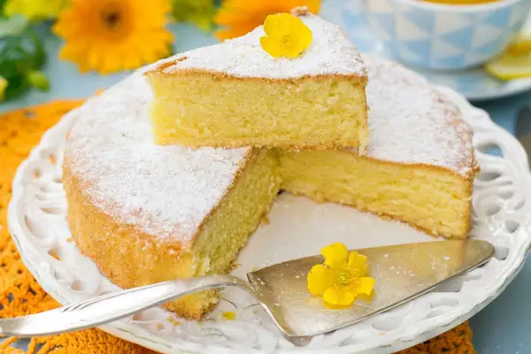 Una fetta di torta paradiso sofficissima, spolverata di zucchero a velo e decorata con un fiore giallo, su un piatto bianco tra fiori arancioni.