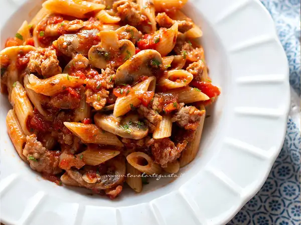 Piatto di pasta alla boscaiola con penne, funghi, salsiccia e pomodoro, guarnito con prezzemolo.