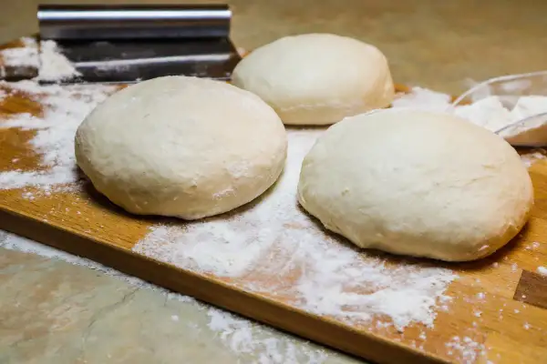 Tre panetti soffici di pizza a lunga lievitazione, pronti per essere stesi su un tagliere di legno infarinato.
