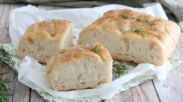 Focaccia senza glutine soffice e dorata, guarnita con rosmarino fresco e piccoli fiori viola, su carta da forno.