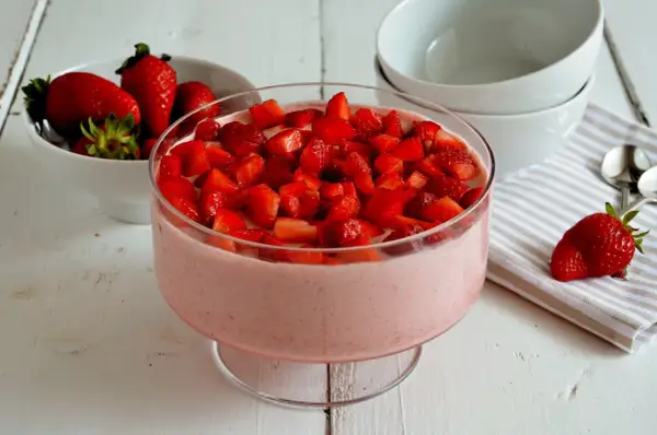Delizioso mousse di fragole in una coppa di vetro, guarnito con fragole fresche a pezzi. Accanto, una ciotola di fragole e ciotole bianche.