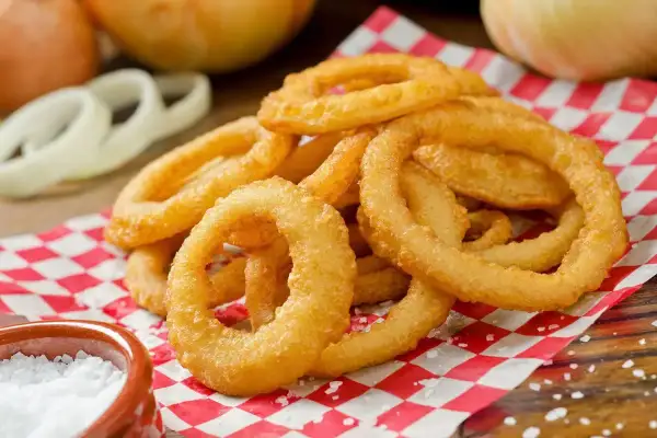 Anelli di cipolla dorati e croccanti, preparati con una perfetta pastella per fritti, serviti su carta a scacchi rossi e bianchi, con sale grosso a lato.