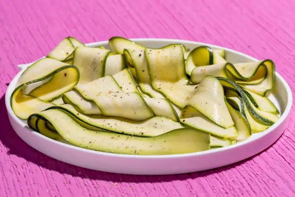Fettine sottili di zucchine marinate crude, condite con pepe nero, disposte a spirale in un piatto bianco su un tavolo rosa acceso.