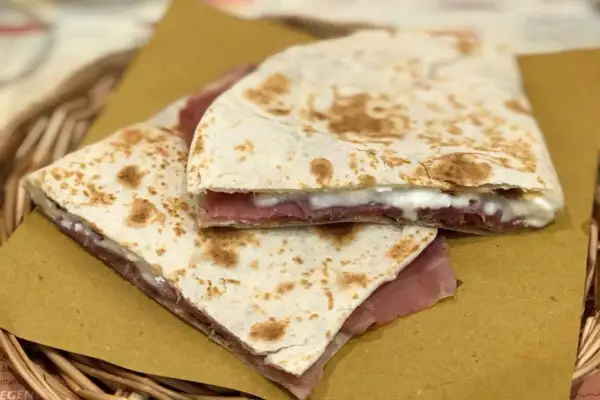 Piadina fatta in casa con prosciutto e formaggio filante, servita su carta gialla in un cestino di vimini.