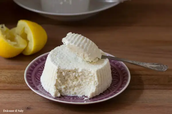 Ricotta fatta in casa su un piatto viola, con limone a fianco. Un cucchiaino preleva una porzione cremosa.