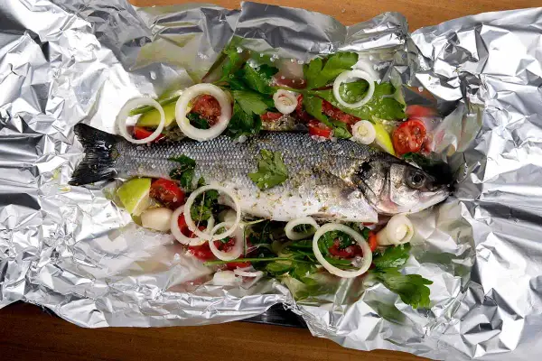 Branzino al cartoccio pronto per la cottura, con pomodorini, cipolla e prezzemolo su foglio d'alluminio.