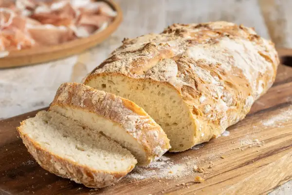 Pane fatto in casa con farina 00, tagliato a fette su un tagliere di legno, con affettati in lontananza.