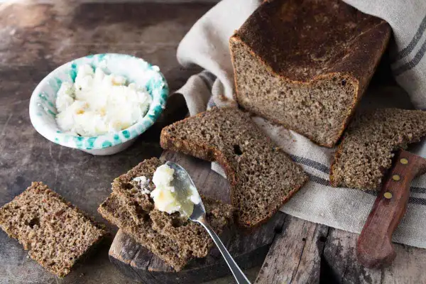Fetta di pane di segale con burro spalmato, accanto a una ciotola di formaggio fresco e un coltello.