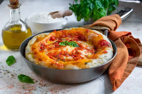 Pizza in padella appena sfornata, con pomodoro, mozzarella filante e basilico fresco. Accanto, olio d'oliva e farina, ingredienti per un'ottima pizza fatta in casa.